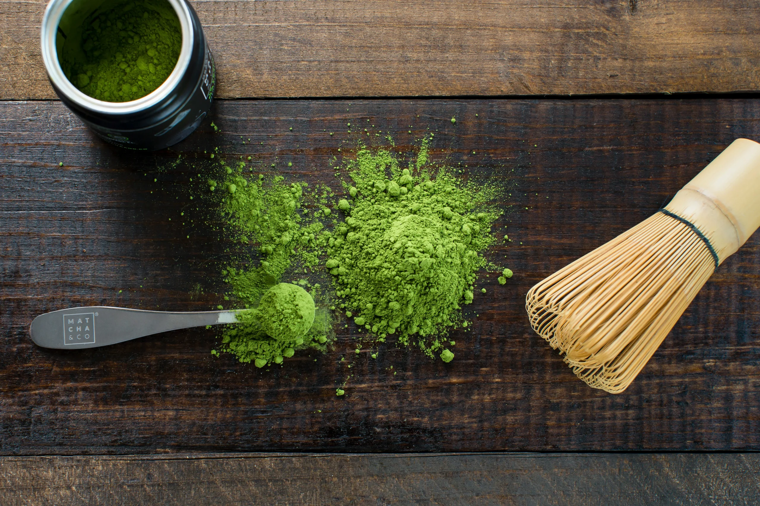 Zielona herbata matcha w proszku marki Matcha & Co, rozsypana na drewnianym blacie. Obok znajduje się tradycyjna bambusowa trzepaczka (chasen) oraz miarka do odmierzania herbaty. Idealna do przygotowania zdrowych napojów, latte i deserów.